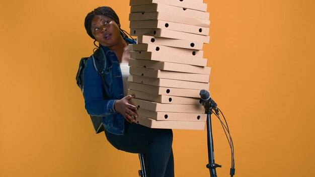 Jeugdige zwarte vrouw die een stapel pizzadozen vasthoudt voor bezorging bij een feestevenement in de buurt. afro-amerikaanse bezorger balanceert voorzichtig enorme bestelling voor voedselbezorging.