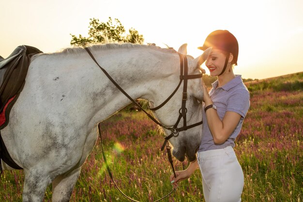 Jockey jong meisje aaien en knuffelen wit paard in avond zonsondergang. zonnevlam