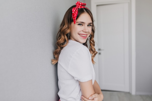 Jocund stijlvolle meisje poseren thuis met een glimlach. indoor foto van prachtig vrouwelijk model draagt rood lint kijkt over de schouder.