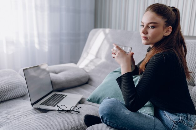 Jong aantrekkelijk meisje dat thuis warme thee drinkt en met laptop werkt. comfort en gezelligheid terwijl u thuis bent. thuiskantoor en thuiswerken. online werkgelegenheid op afstand.