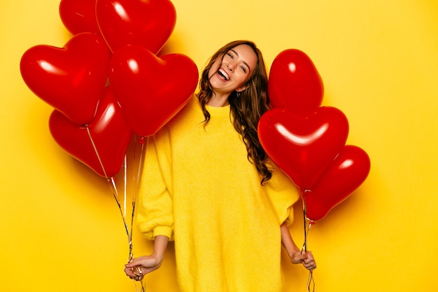 Jong aantrekkelijk meisje met lang krullend haar, in gele sweater die rode luchtballons houden