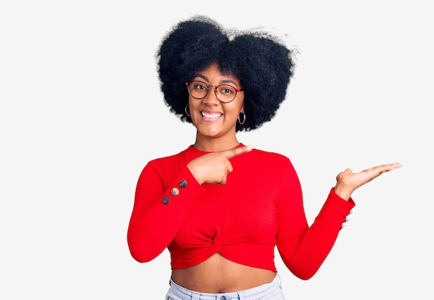 Gratis foto jong afrikaans amerikaans meisje met casual kleding en bril verbaasd en glimlachend naar de camera terwijl ze met de hand presenteert en met de vinger wijst.