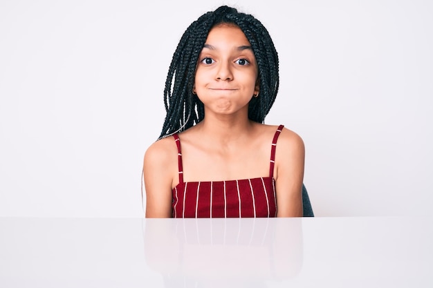 Gratis foto jong afrikaans amerikaans meisjeskind met vlechten die casual kleding dragen zittend op de tafel puffende wangen met een grappig gezicht. mond opgeblazen met lucht, gekke uitdrukking.