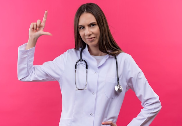 jong artsenmeisje dat stethoscoop medische toga draagt die grootte op geïsoleerde roze muur toont