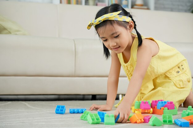 Jong Aziatisch meisje dat op vloer thuis knielt en met kleurrijke bouwstenen speelt