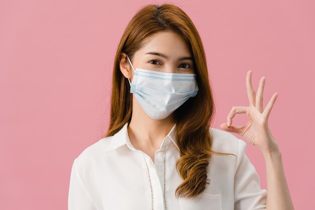 Jong Azië meisje met medisch gezichtsmasker gebaren ok teken met gekleed in casual doek en kijk naar camera geïsoleerd op roze achtergrond.