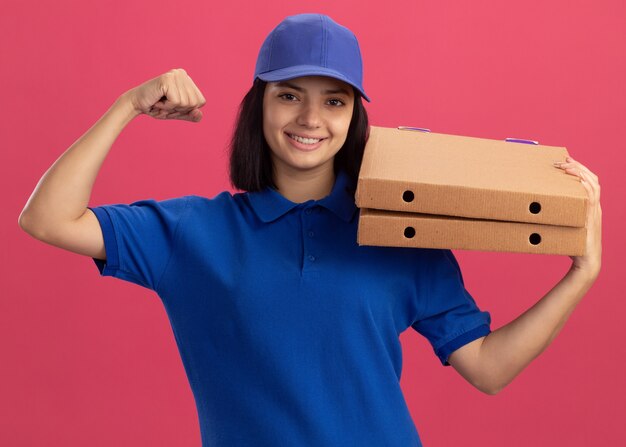 Jong bezorgermeisje in blauw uniform en pet met pizzadozen die vuist opheffen als een winnaar die vrolijk over roze muur glimlacht