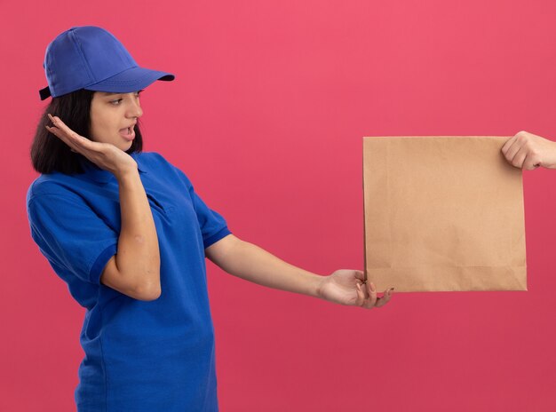 Jong bezorgmeisje in blauw uniform en pet die zich opgewonden voelen terwijl ze een papieren pakket ontvangt dat zich over de roze muur bevindt