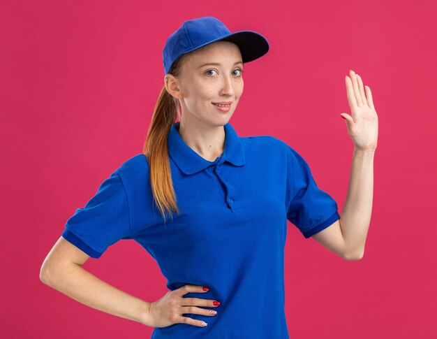 Jong bezorgmeisje in blauw uniform en pet glimlachend zelfverzekerd met open arm die over roze muur staat