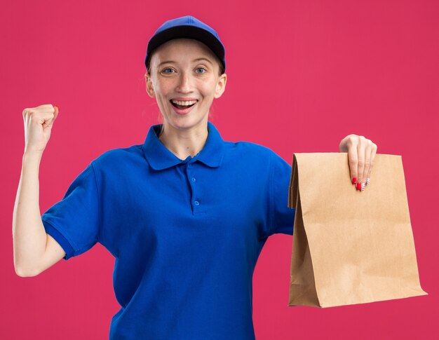 Jong bezorgmeisje in blauw uniform en pet met papieren pakketje blij en opgewonden, gebalde vuist over roze muur