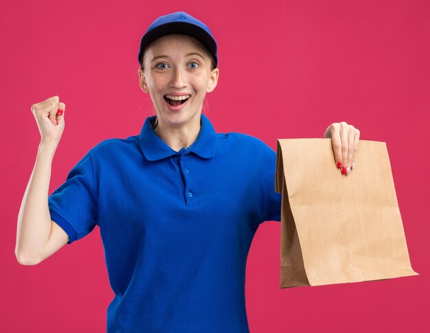 Jong bezorgmeisje in blauw uniform en pet met papieren pakketje blij en opgewonden gebalde vuist