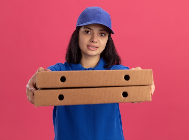 Jong bezorgmeisje in blauw uniform en pet met pizzadozen die zich uitstrekken op zoek naar zelfverzekerd staande over roze muur