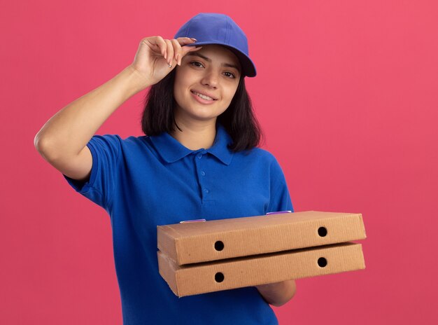 Jong bezorgmeisje in blauw uniform en pet met pizzadozen glimlachend zelfverzekerd aanraken van haar pet staande over roze muur