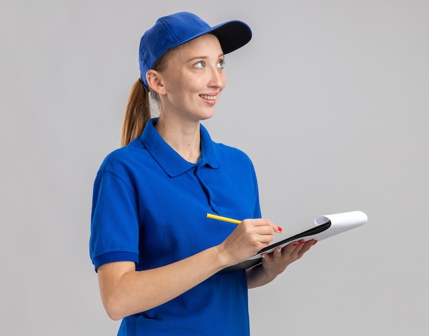 Jong bezorgmeisje in blauw uniform en pet met potlood en klembord met blanco pagina's opzij kijkend met een glimlach op het gezicht