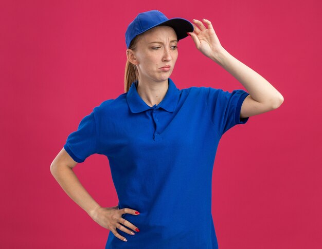 Jong bezorgmeisje in blauw uniform en pet opzij kijkend met een sceptische uitdrukking op het gezicht dat de pet aanraakt