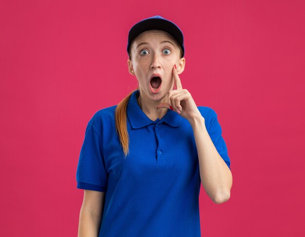 Jong bezorgmeisje in blauw uniform en pet verbaasd en verrast staande over roze muur