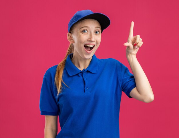 Jong bezorgmeisje in blauw uniform en pet verrast en blij dat wijsvinger een nieuw idee heeft dat over een roze muur staat