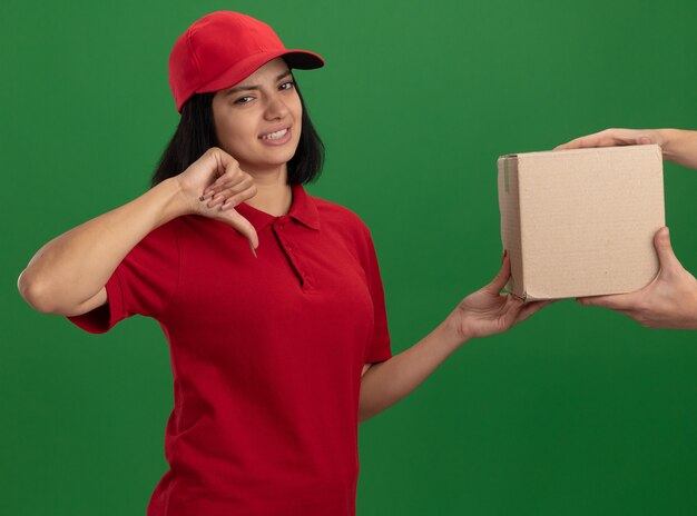 Jong bezorgmeisje in rood uniform en pet met kartonnen doos naar een klant die ontevreden kijkt met duimen naar beneden staande over groene muur