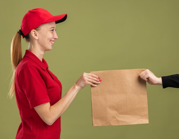 Jong bezorgmeisje in rood uniform en pet met papieren pakket en geeft het aan een klant die zelfverzekerd over groene muur glimlacht