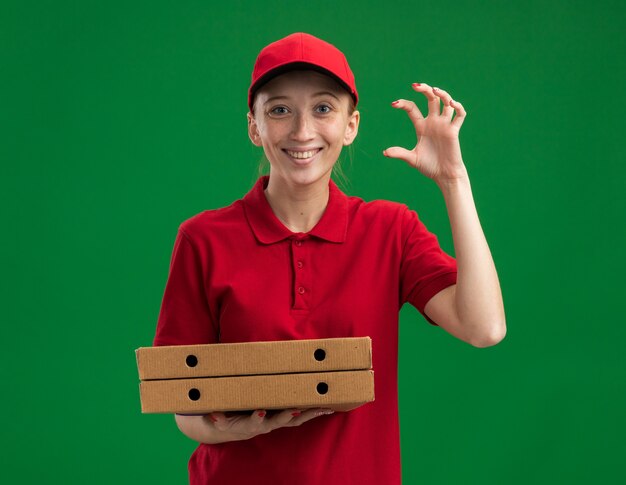 Jong bezorgmeisje in rood uniform en pet met pizzadozen die een klein gebaar maken met vingers die vrolijk glimlachen over de groene muur