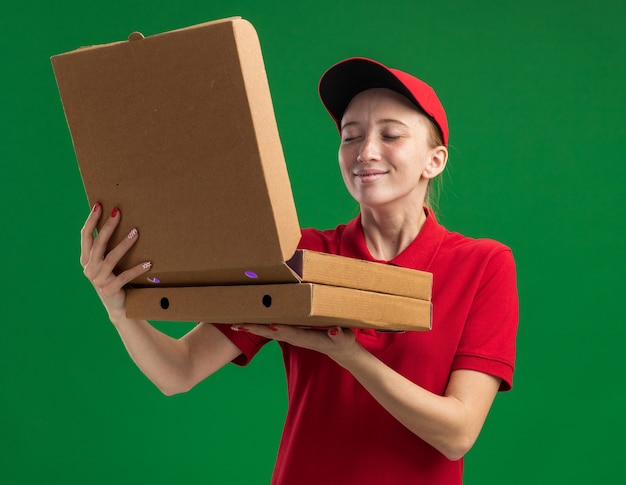 Jong bezorgmeisje in rood uniform en pet met pizzadozen die een van de dozen openen die een aangenaam aroma inademen dat over de groene muur staat