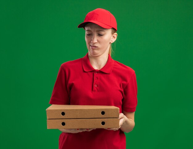 Jong bezorgmeisje in rood uniform en pet met pizzadozen die verward en ontevreden naar ze kijken terwijl ze over de groene muur staan