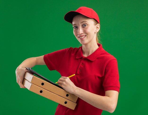 Jong bezorgmeisje in rood uniform en pet met pizzadozen en klembord met blanco pagina's met potlood glimlachend zelfverzekerd over groene muur