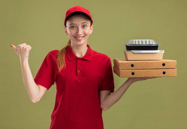 Jong bezorgmeisje in rood uniform en pet met pizzadozen en voedselpakketten glimlachend zelfverzekerd wijzend naar de zijkant over groene muur