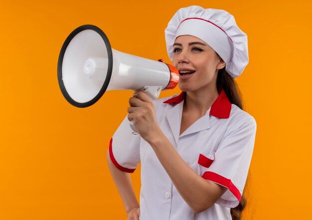 Jong blij Kaukasisch kokmeisje in eenvormige chef-kok schreeuwt door luide spreker die op oranje muur met exemplaarruimte wordt geïsoleerd