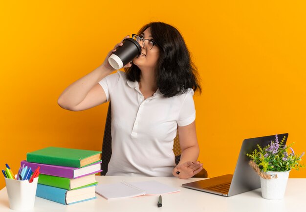 Jong blij vrij Kaukasisch schoolmeisje met bril zit aan bureau met schoolgereedschap drinkt kopje koffie opzoeken op oranje met kopie ruimte