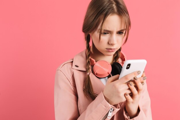 Jong boos meisje met twee vlechten in leren jas met koptelefoon die helaas mobiele telefoon gebruikt terwijl ze tijd doorbrengt op roze achtergrond