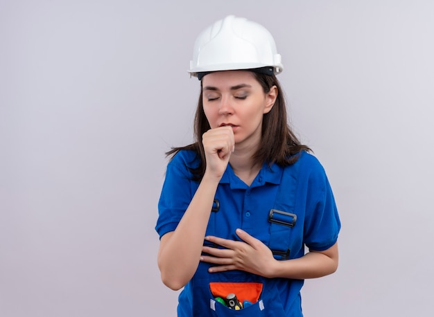 Jong bouwersmeisje met witte veiligheidshelm en blauw uniform beweert te hoesten op geïsoleerde witte achtergrond met exemplaarruimte