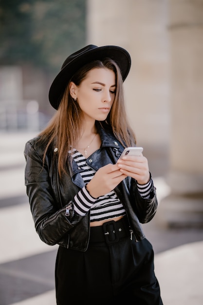 Jong bruinharige meisje in een leren jas, zwarte hoed op de stadspromenade en speelt op de mobiele telefoon