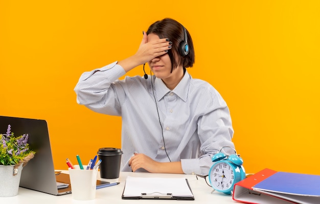 Jong call centreermeisje die hoofdtelefoonszitting bij bureau met uitrustingsstukken dragen die ogen met hand sluiten die op oranje achtergrond worden geïsoleerd