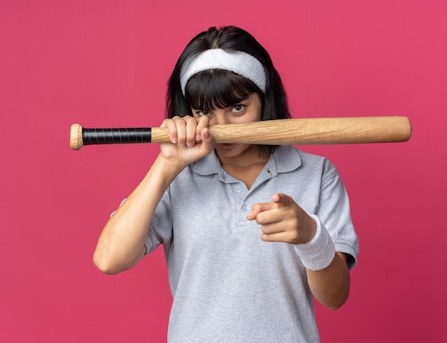 Jong fitness meisje met hoofdband met honkbalknuppel kijkend naar camera met zelfverzekerde glimlach wijzend met wijsvinger naar camera staande over roze achtergrond