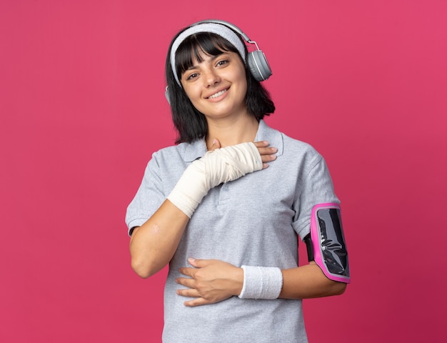 Jong fitness meisje met hoofdband met koptelefoon en armband voor smartphone kijkend naar camera glimlachend zelfverzekerd over roze achtergrond