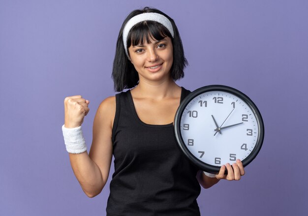 Jong fitness meisje met hoofdband met wandklok kijkend naar camera, blij en zelfverzekerd gebalde vuist over blauwe achtergrond