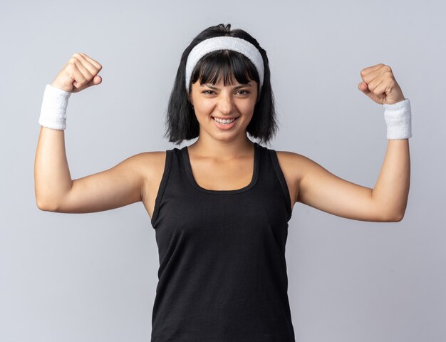 Jong fitnessmeisje dat een hoofdband draagt en naar de camera kijkt, gespannen en zelfverzekerd met het opheffen van vuisten die op een witte achtergrond staan