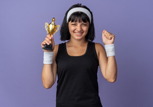 Jong fitnessmeisje met een hoofdband die een trofee vasthoudt, blij en opgewonden met een vuist die zich verheugt over haar succes dat over blauw staat