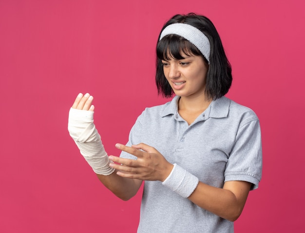 Jong fitnessmeisje met een hoofdband die haar verbonden hand aanraakt en er onwel uitziet en pijn voelt