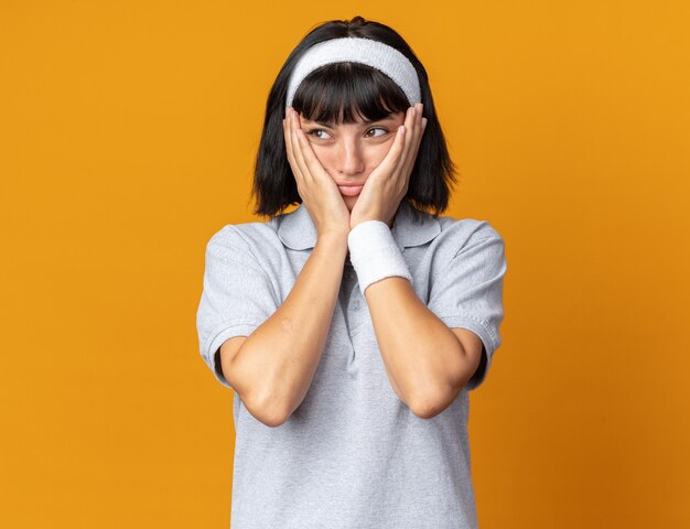 Jong fitnessmeisje met een hoofdband die verward en bezorgd opzij kijkt met handen op haar wangen die over oranje staan