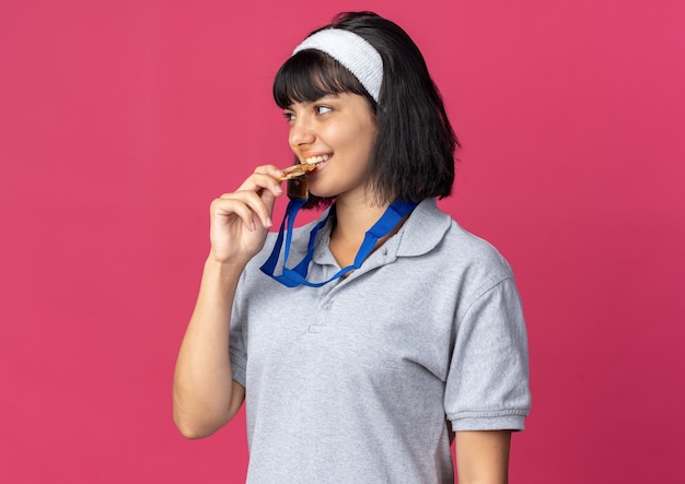 Jong fitnessmeisje met een hoofdband met een gouden medaille om de nek die erop bijt als een kampioen die over een roze achtergrond staat