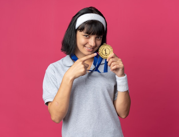 Jong fitnessmeisje met een hoofdband met een gouden medaille om de nek, wijzend met de wijsvinger erop glimlachend vrolijk over een roze achtergrond