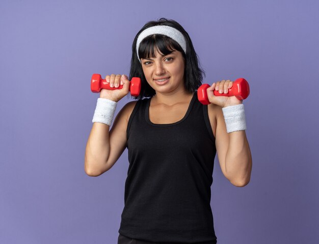 Jong fitnessmeisje met een hoofdband met halters die oefeningen doen die er verward uitzien