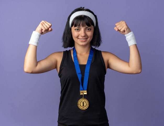 Jong fitnessmeisje met hoofdband met gouden medaille om nek die vuisten opheft, blij en zelfverzekerd poserend als een winnaar die over een blauwe achtergrond staat