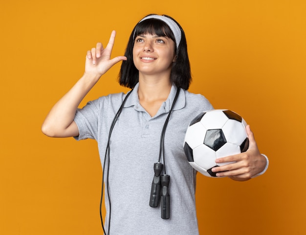 Jong fitnessmeisje met hoofdband met springtouw om nek met voetbal en glimlachend omhoog kijkend met wijsvinger