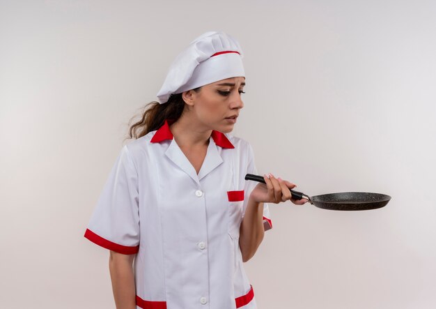 Jong geïrriteerd Kaukasisch kokmeisje in eenvormige chef-kok houdt en bekijkt koekenpan die op witte achtergrond met exemplaarruimte wordt geïsoleerd