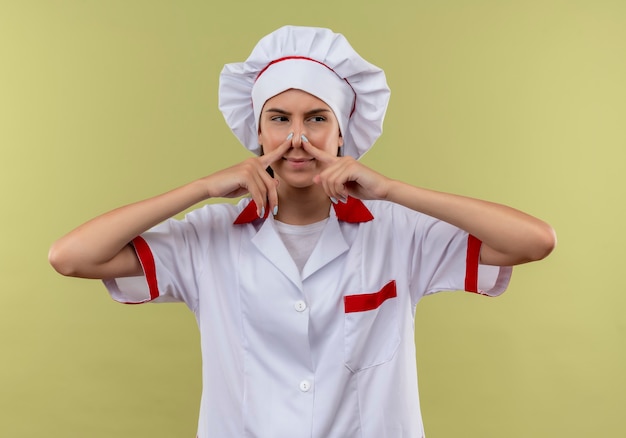 Gratis foto jong geïrriteerd kaukasisch kokmeisje in eenvormige chef-kok sluit neus en bekijkt kant op groen met exemplaarruimte