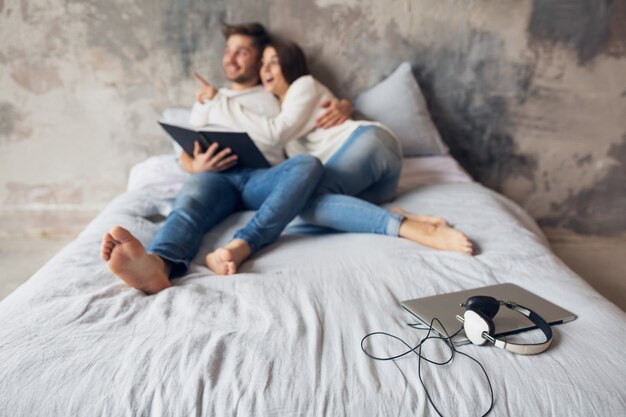 Jong gelukkig lachend paar zittend op bed thuis in casual outfit leesboek dragen jeans, man en vrouw benen romantische stemming, laptop