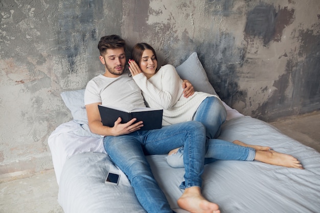 Jong gelukkig lachend paar zittend op bed thuis in casual outfit leesboek dragen jeans, man en vrouw romantische tijd samen doorbrengen